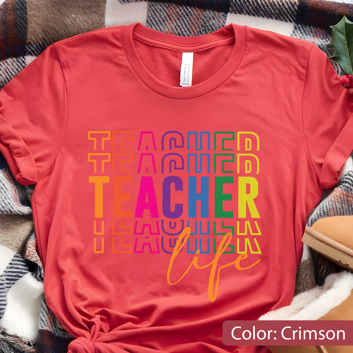 May include: A crimson short-sleeved t-shirt featuring the word TEACHER repeated in a stacked, colorful, block-letter design. Below the stacked text, the word life is printed in a yellow cursive script. The shirt has a round neckline.