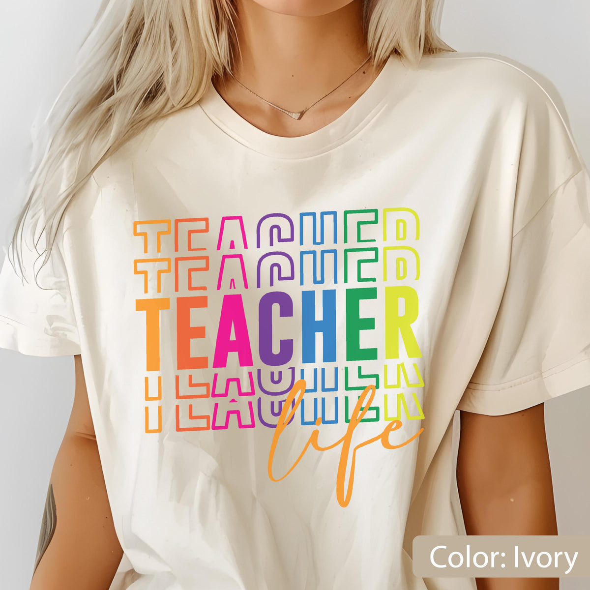 May include: An ivory short-sleeved t-shirt featuring a multi-colored graphic print. The design displays the word TEACHER repeated in stacked, outlined block letters in rainbow colors, with the word life written in orange cursive script below.