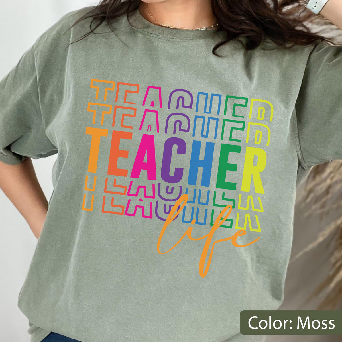 May include: A moss green short-sleeve t-shirt featuring the word TEACHER printed in large, colorful, stacked block letters. Below the main text, the word life is written in a cursive orange script. The design uses a vibrant rainbow color palette.