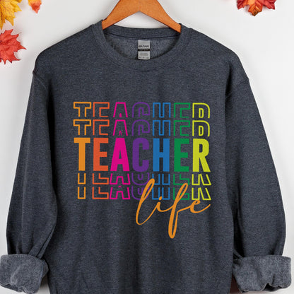 May include: A dark gray sweatshirt with the word "TEACHER" in a rainbow-colored, stacked design. The word "life" is written in an orange script font below the word "TEACHER". The sweatshirt is hanging on a wooden hanger.