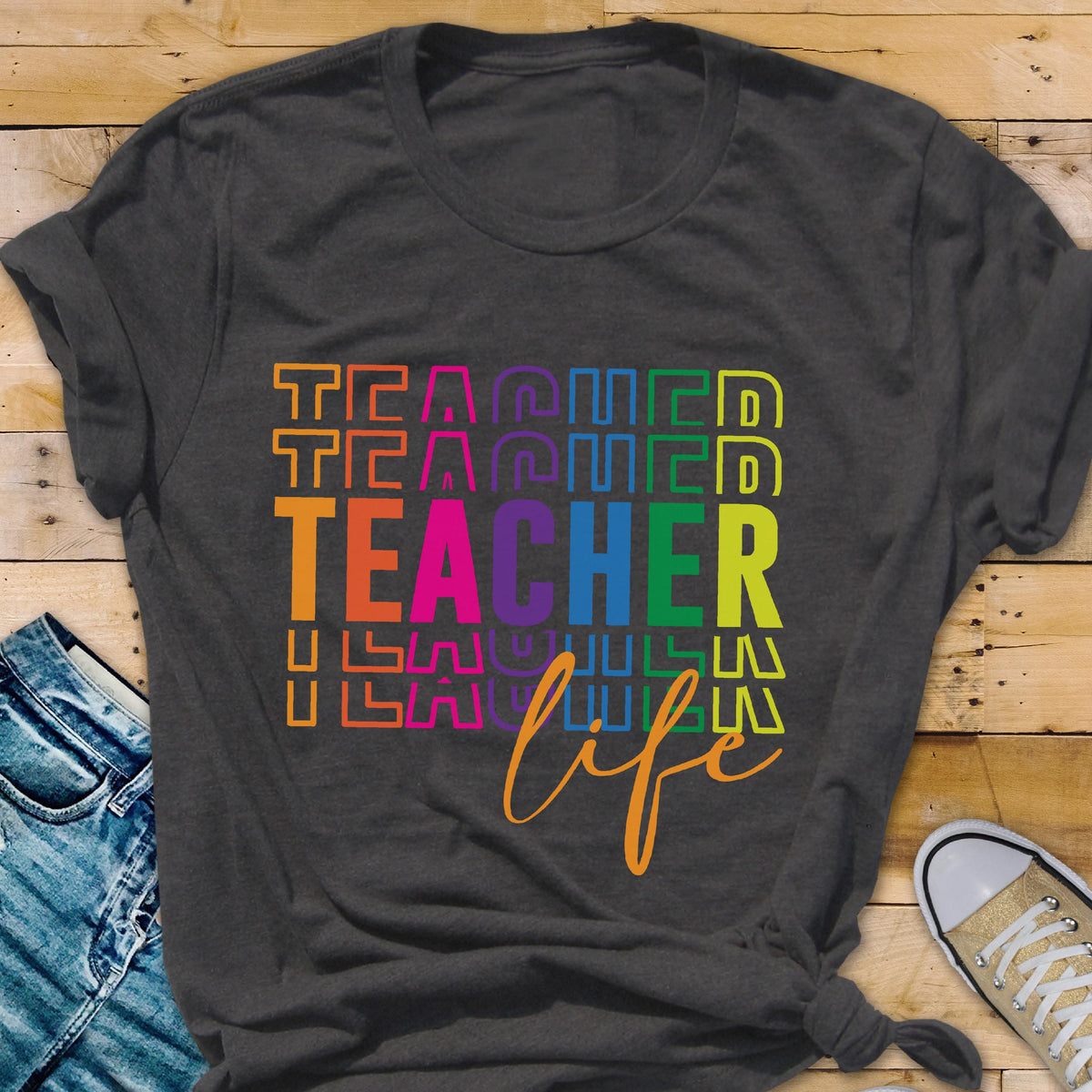 May include: Dark gray t-shirt with the word "TEACHER" in large, colorful letters. The word "TEACHER" is stacked vertically with the word "life" in a cursive font below it. The shirt is on a wooden surface.