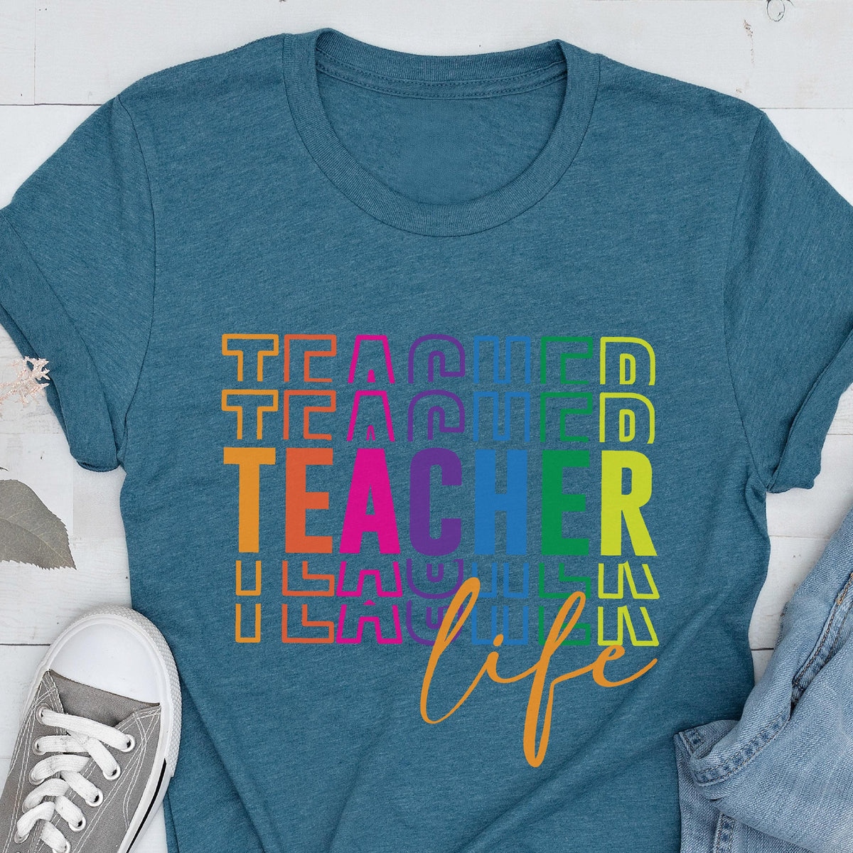 May include: Teal t-shirt with the word "TEACHER" in colorful stacked letters and the word "life" in cursive. A gray canvas sneaker is visible in the lower left corner. The shirt is made of a soft material.