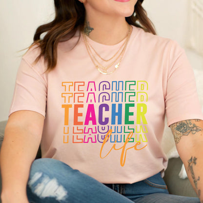 May include: A light pink t-shirt with the word "TEACHER" in large, rainbow-colored letters. The word "TEACHER" is stacked multiple times, with the word "life" in cursive below. The shirt is a crew neck.