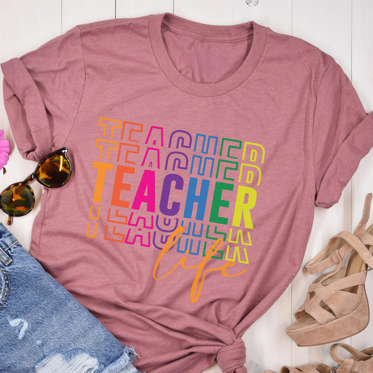 May include: A dusty rose t-shirt with the word "TEACHER" in a rainbow gradient design. The word "life" is written in a cursive font below the word "TEACHER". Sunglasses and denim shorts are visible in the image.