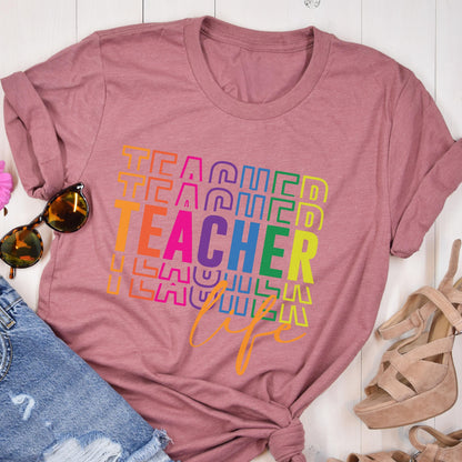 May include: A dusty rose t-shirt with the word "TEACHER" in a rainbow gradient design. The word "life" is written in a cursive font below the word "TEACHER". Sunglasses and denim shorts are visible in the image.