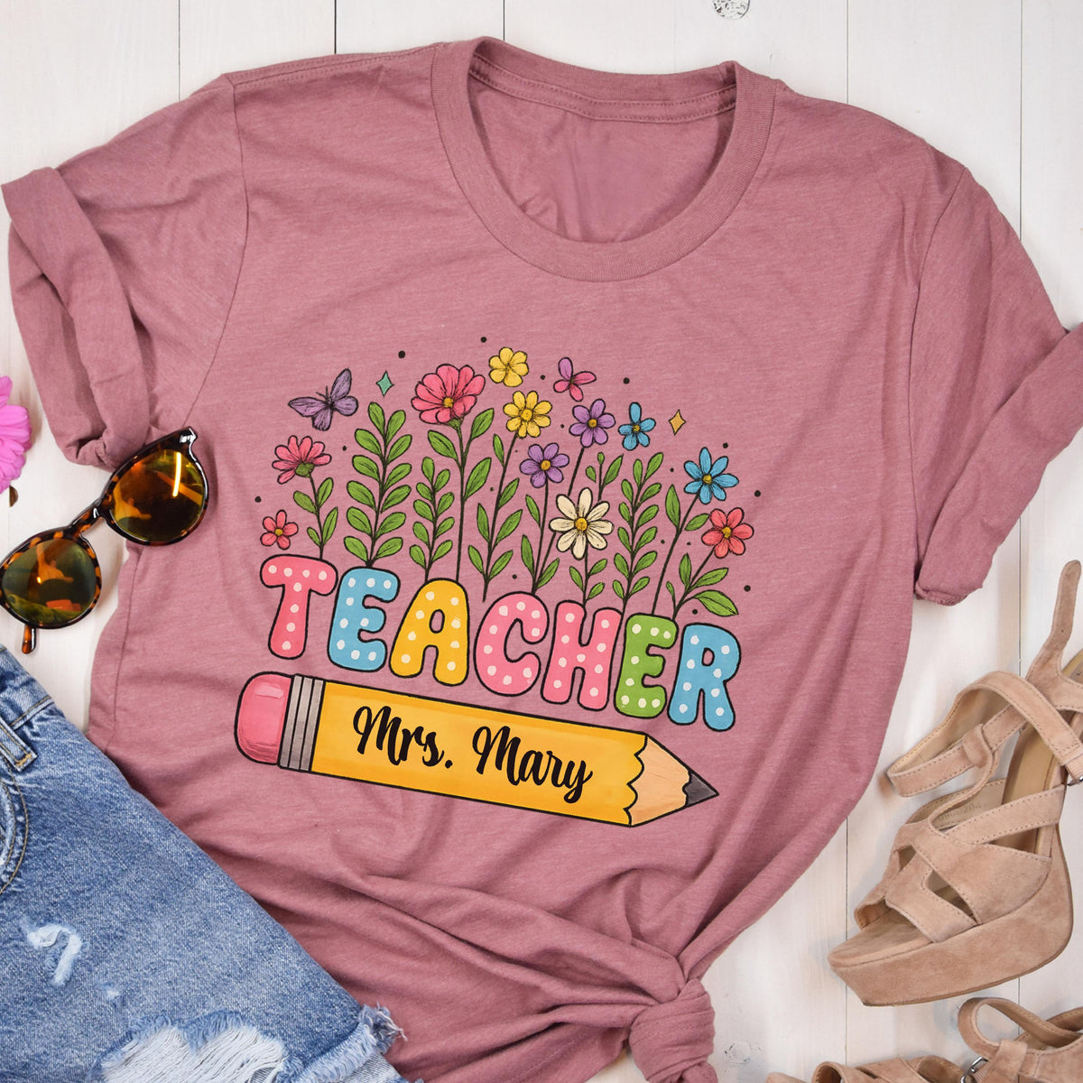 May include: A mauve-colored t-shirt featuring the word "TEACHER" in colorful bubble letters, with a pencil graphic below and the name "Mrs. Mary". The design includes a floral arrangement with butterflies.