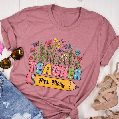 May include: A mauve-colored t-shirt featuring the word "TEACHER" in colorful bubble letters, with a pencil graphic below and the name "Mrs. Mary". The design includes a floral arrangement with butterflies.