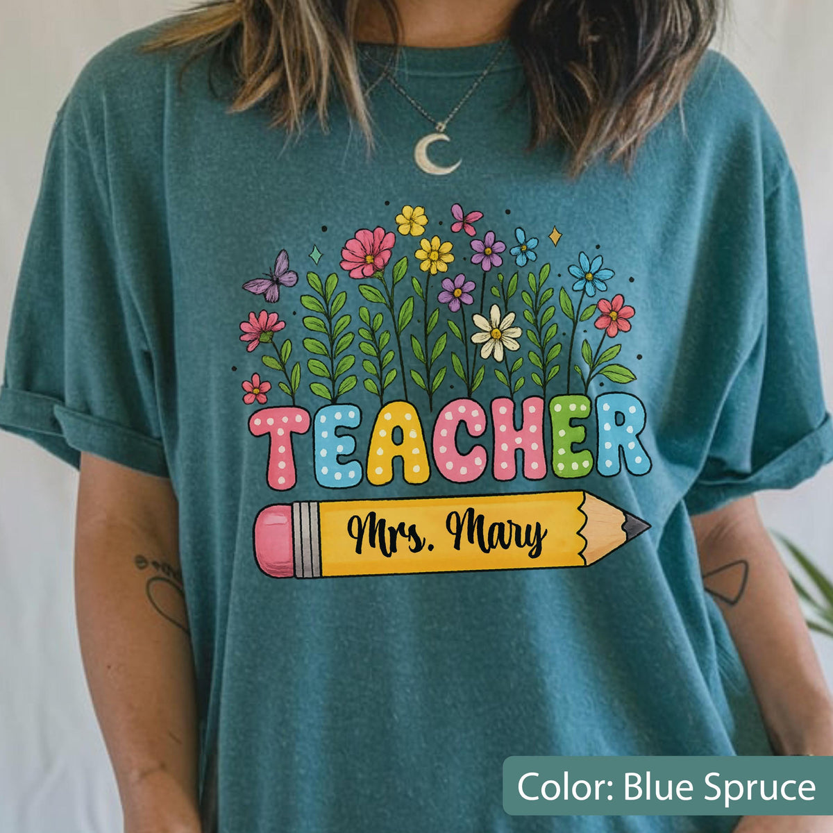 May include: Blue spruce t-shirt with a colorful floral design and the word "TEACHER" in large, playful letters. A yellow pencil with the name "Mrs. Mary" is featured below the word.
