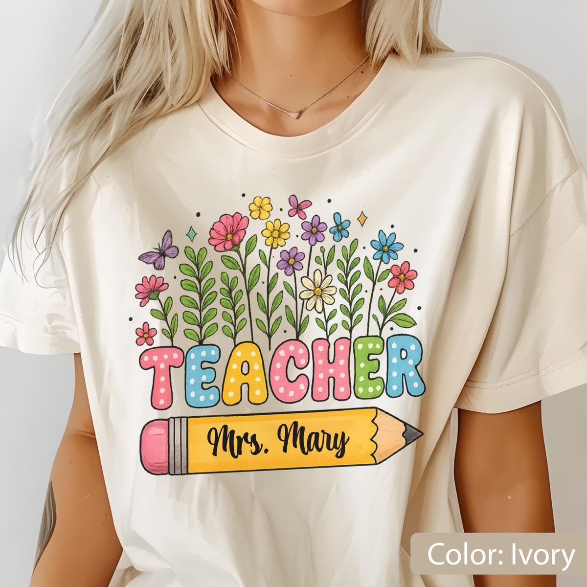 May include: Ivory-colored t-shirt featuring a colorful floral design with the word "TEACHER" in large, multi-colored letters. Below the word is a yellow pencil with the name "Mrs. Mary". The shirt is a casual style, perfect for teachers.