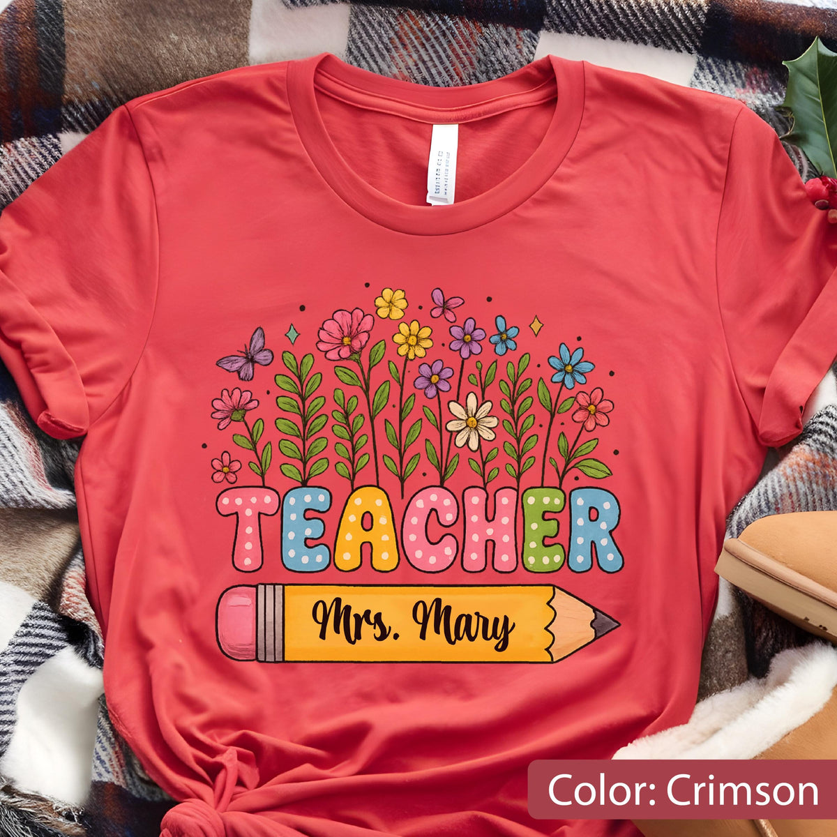 May include: Crimson-colored t-shirt featuring a colorful floral design and the word "TEACHER" in bubble letters. Below the word is a yellow pencil with the name "Mrs. Mary". The shirt is made of soft material.