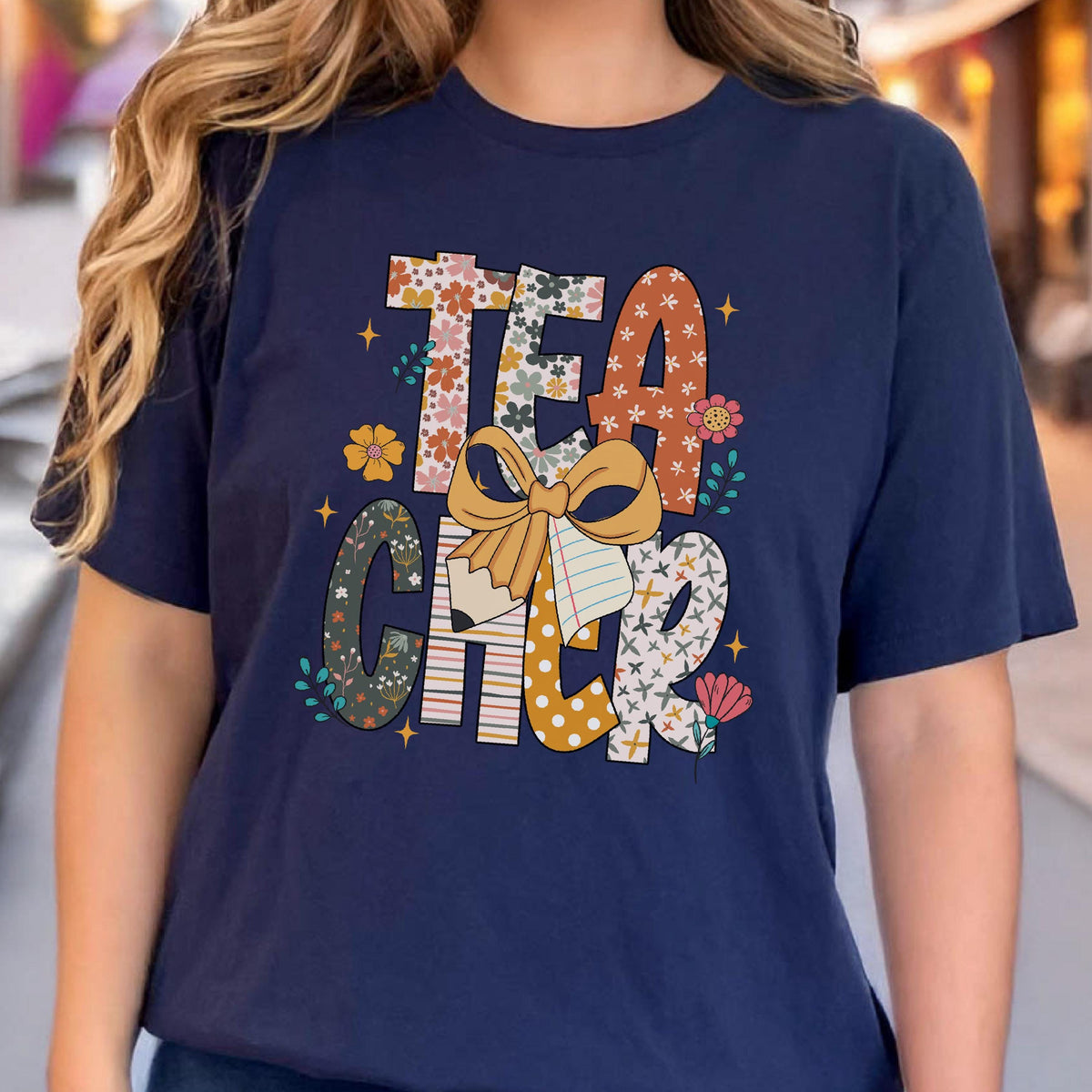 May include: Navy blue t-shirt with the word "TEACHER" in a colorful, floral design. The letters are filled with various patterns and colors, including flowers, polka dots, and stripes. A large bow and paper add to the design.