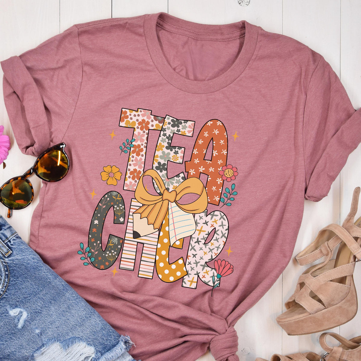 May include: A dusty rose-colored t-shirt with the word "TEACHER" in a colorful, floral design. The letters are filled with patterns, flowers, and a pencil with a bow. Sunglasses, denim shorts, and platform sandals are visible.