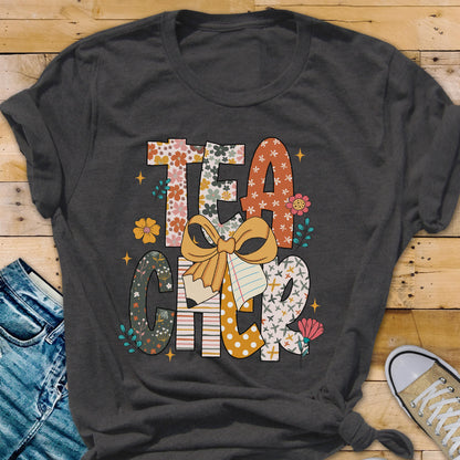 May include: Dark gray t-shirt with the word "TEACHER" in a colorful, floral design. The letters are decorated with flowers, patterns, and a bow. A pencil and notebook are also featured in the design. The shirt is folded on a wooden surface.