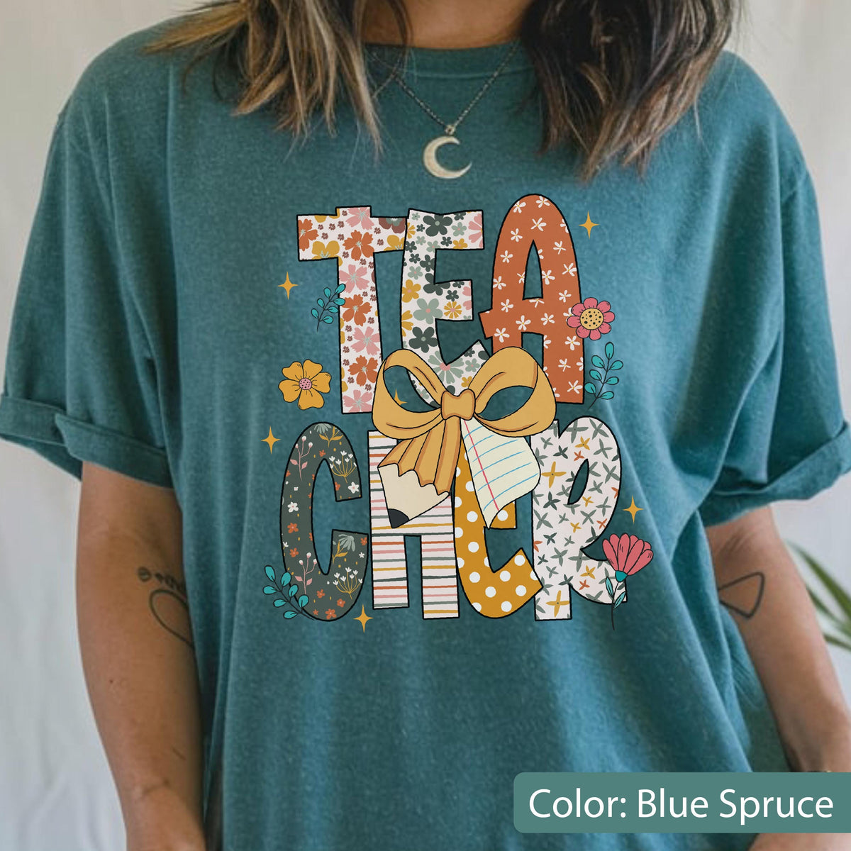 May include: Blue spruce t-shirt with the word "TEACHER" in colorful, patterned letters. The design includes a pencil, a bow, and floral accents. The shirt is a comfortable, casual style.