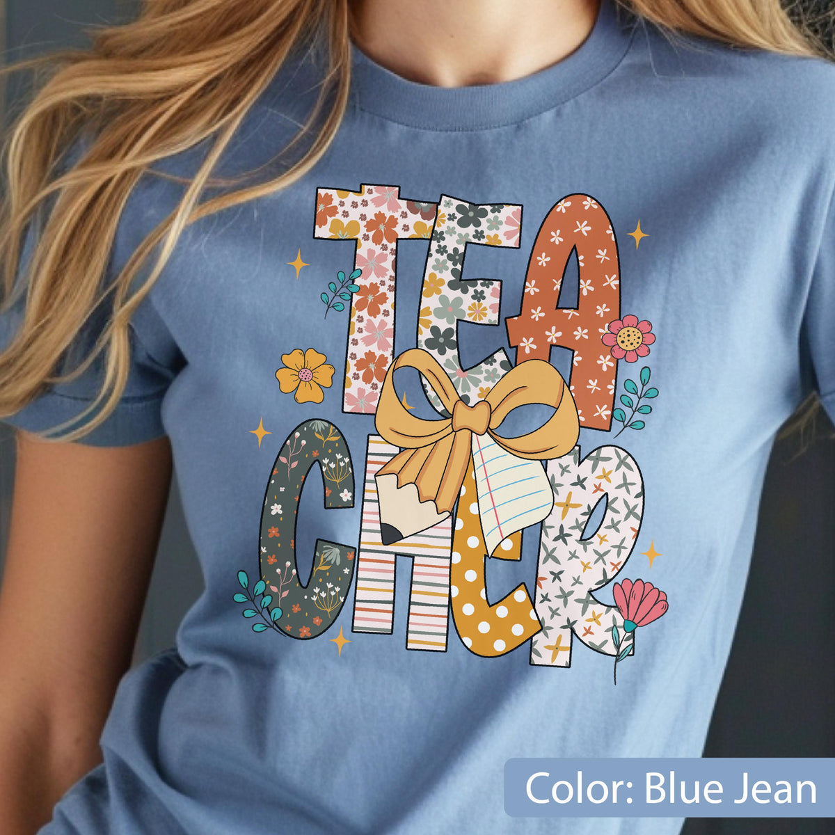 May include: A light blue t-shirt with the word "TEACHER" in a colorful, floral design. The letters are filled with patterns, including flowers, stripes, and polka dots. A large bow and pencil graphic are in the center.