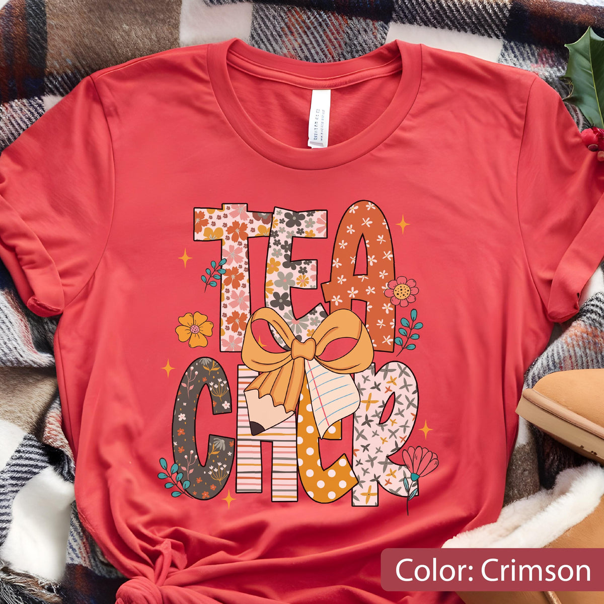 May include: Crimson-colored t-shirt with the word "TEACHER" in a floral and patterned design. The design includes a pencil, a bow, and flowers. The shirt is made of a soft material.