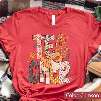 May include: Crimson-colored t-shirt with the word "TEACHER" in a floral and patterned design. The design includes a pencil, a bow, and flowers. The shirt is made of a soft material.