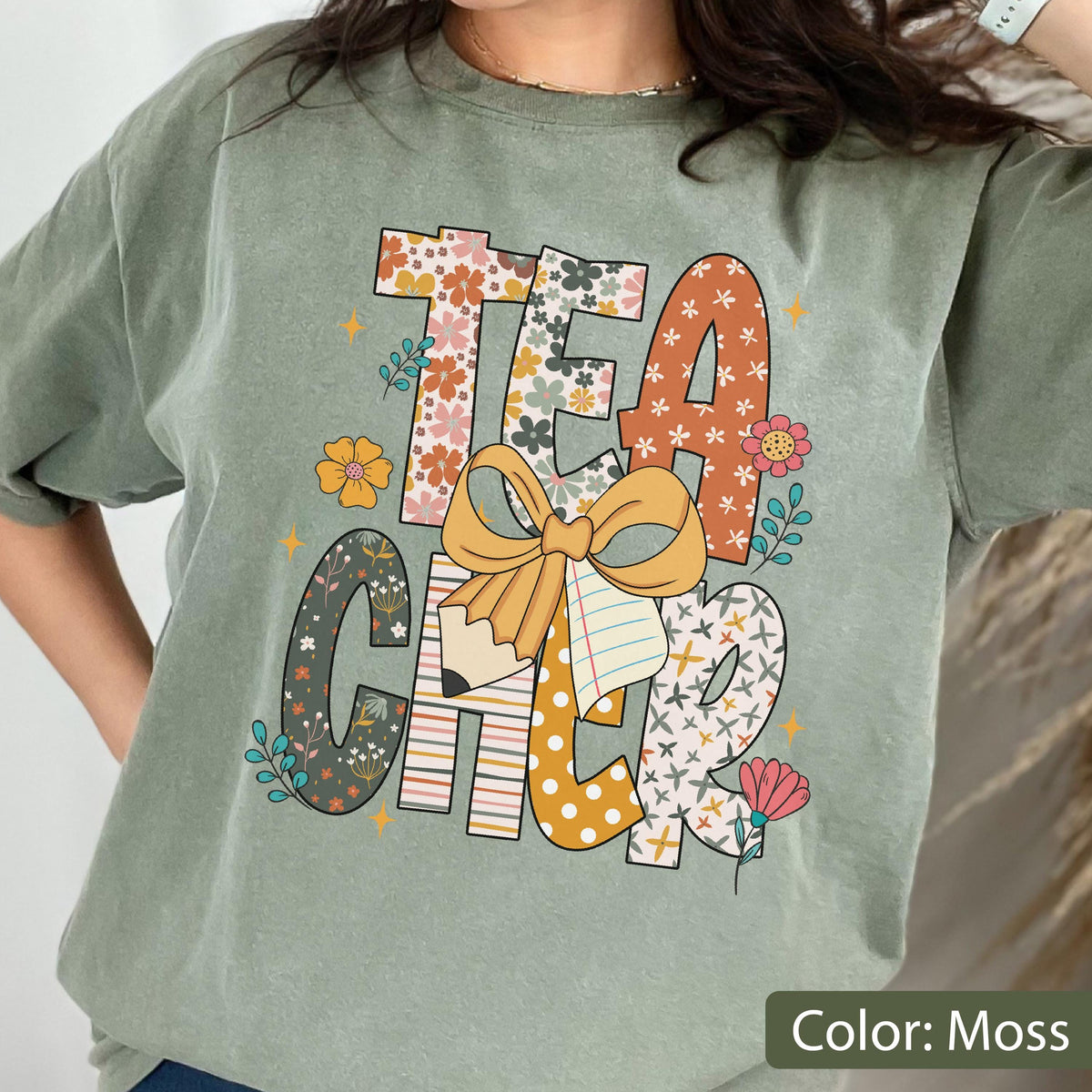 May include: A moss green t-shirt with the word "TEACHER" in a colorful, floral design. The letters are decorated with flowers, bows, and a pencil. The shirt is a comfortable, casual style.