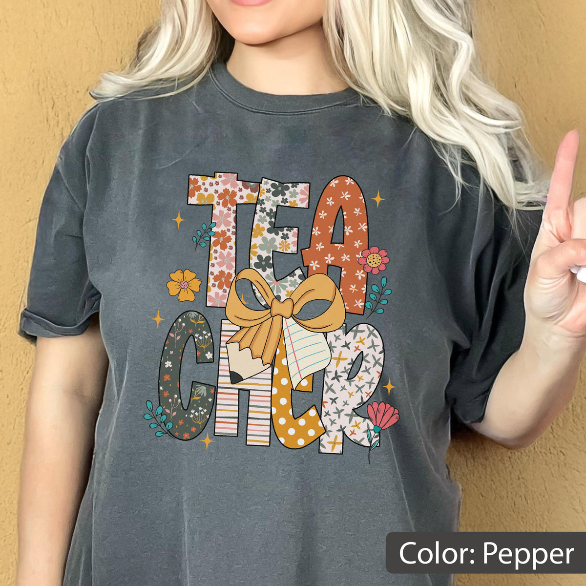 May include: Pepper-colored t-shirt with the word "TEACHER" in a colorful, floral design. The letters are adorned with flowers, bows, and a pencil. The shirt has a vintage, washed look.