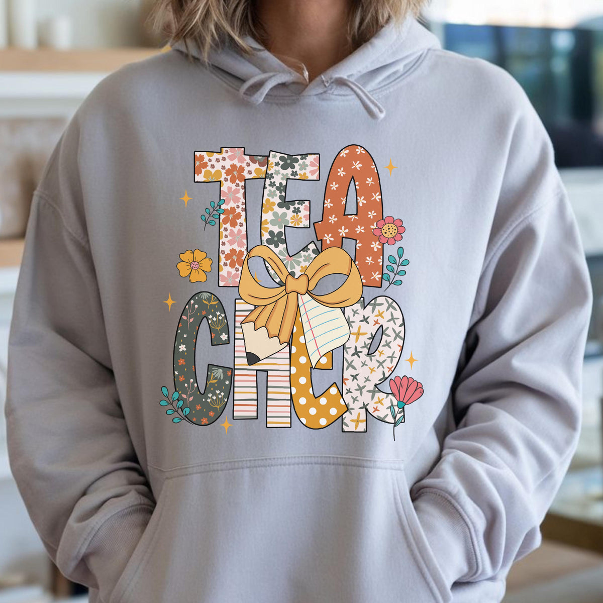 May include: A light gray hoodie with the word "TEACHER" in colorful, patterned letters. The design includes floral patterns, a bow, a pencil, and a notepad. The hoodie has a front pocket and a drawstring hood.