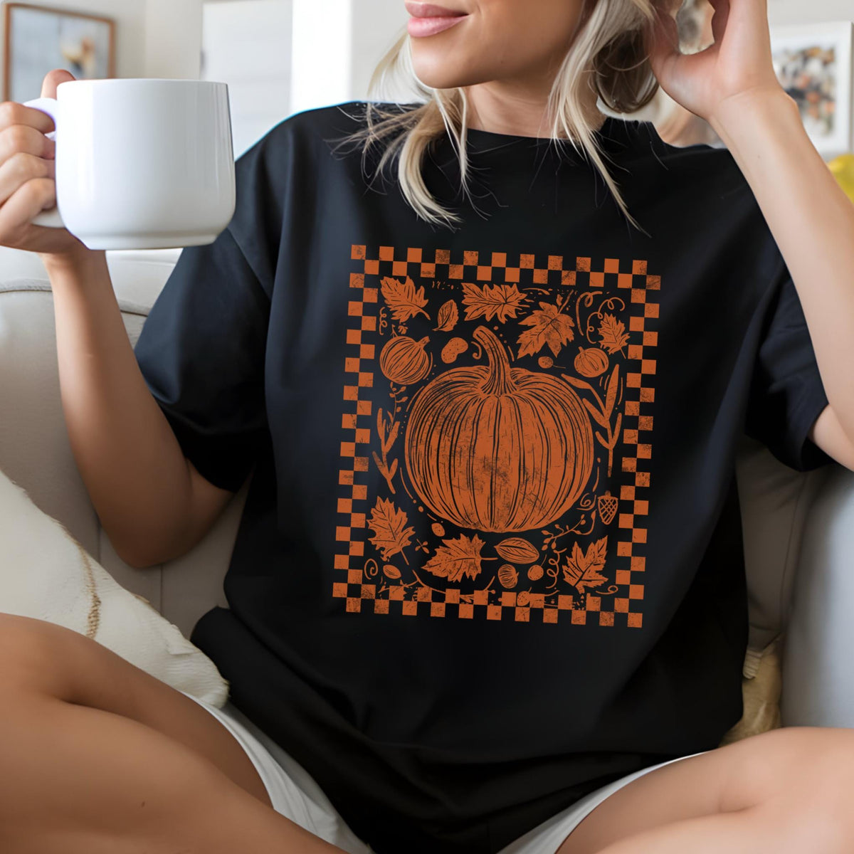 May include: Black t-shirt featuring an orange pumpkin design within a checkered border. The graphic includes leaves and other fall elements. The person is holding a white mug.