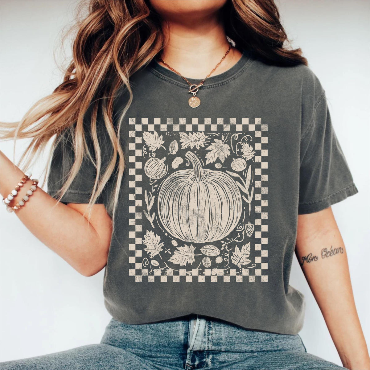 May include: Dark grey t-shirt featuring a cream-colored pumpkin design within a checkered border. The design includes leaves, gourds, and other fall elements. The shirt is a comfortable, casual style.