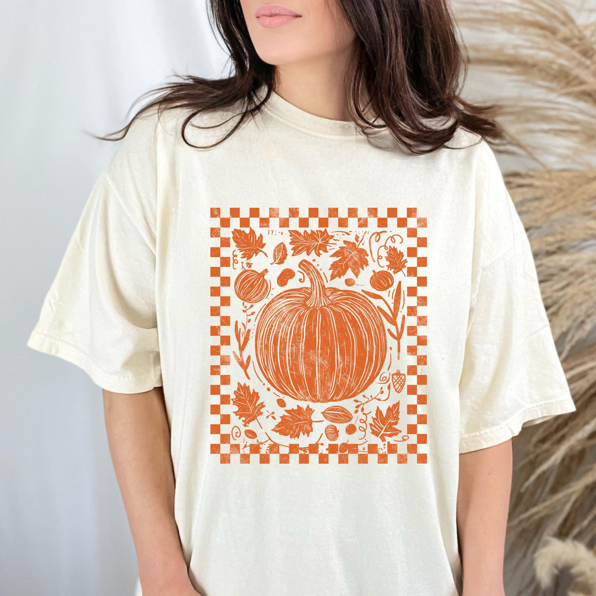 May include: Cream-colored t-shirt featuring an orange pumpkin design with leaves and a checkered border. The graphic is centered on the shirt, creating a fall-themed aesthetic. The shirt has a relaxed fit.