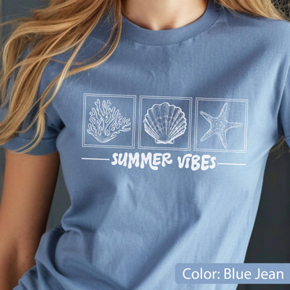 May include: Blue jean-colored t-shirt featuring a white graphic of a coral, seashell, and starfish in square boxes. The words "SUMMER VIBES" are printed below the images. The shirt has short sleeves.