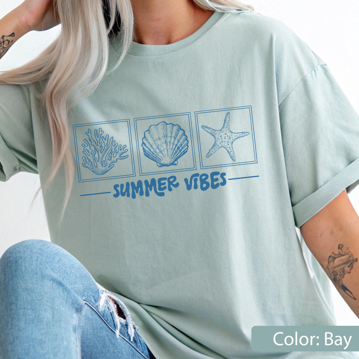 May include: A light green t-shirt with a blue graphic of coral, a seashell, and a starfish in square frames. The words "SUMMER VIBES" are printed below the images. The shirt is a solid color and has rolled-up sleeves.