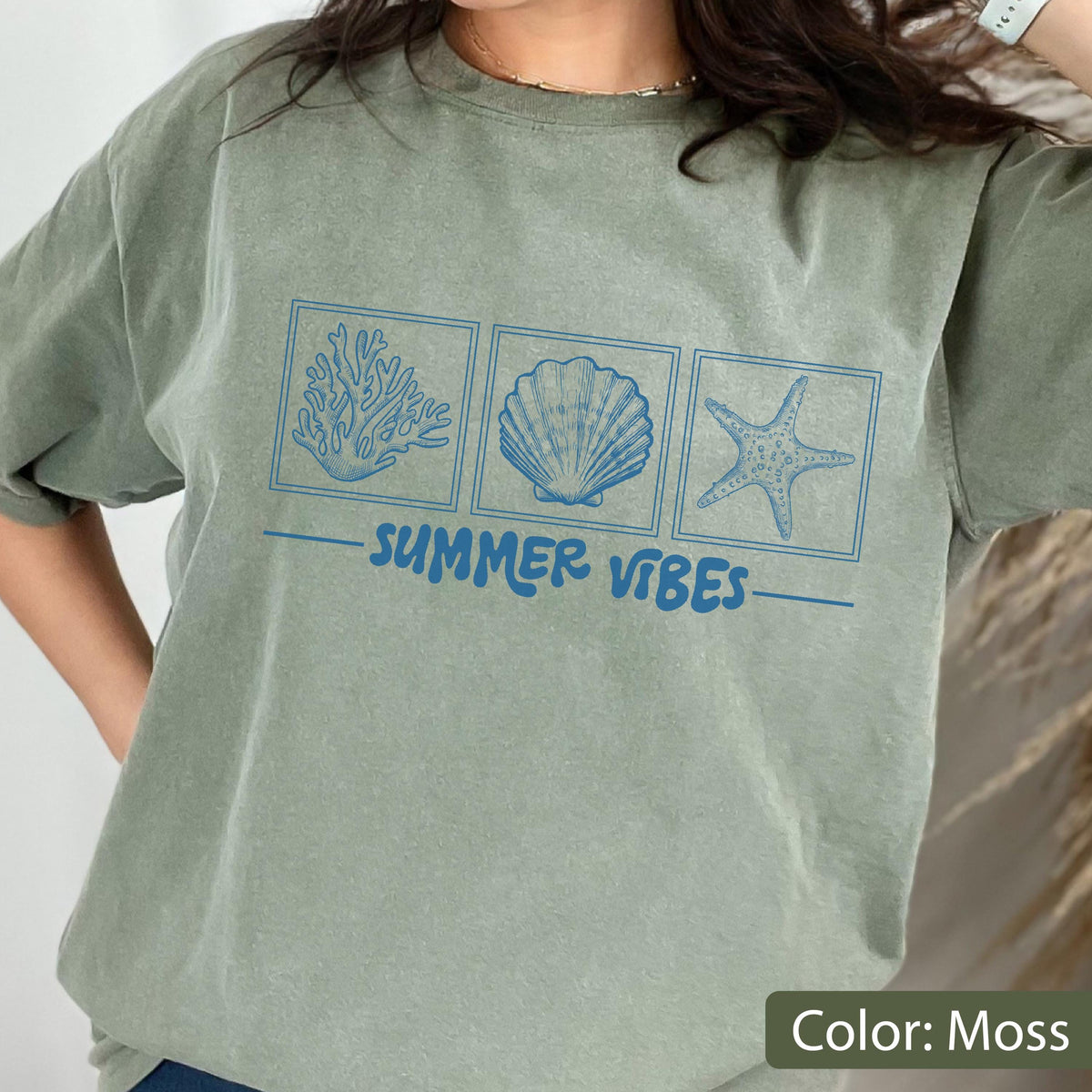 May include: Moss green t-shirt with a blue graphic of coral, a seashell, and a starfish. The words "SUMMER VIBES" are printed in blue below the images. The shirt is a relaxed fit.