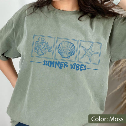 May include: Moss green t-shirt with a blue graphic of coral, a seashell, and a starfish. The words "SUMMER VIBES" are printed in blue below the images. The shirt is a relaxed fit.