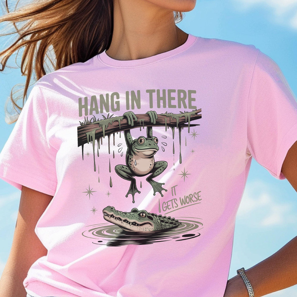 May include: A light pink t-shirt featuring a humorous graphic. The design includes a frog hanging from a branch above an alligator in water, with the text "HANG IN THERE" and "IT GETS WORSE".