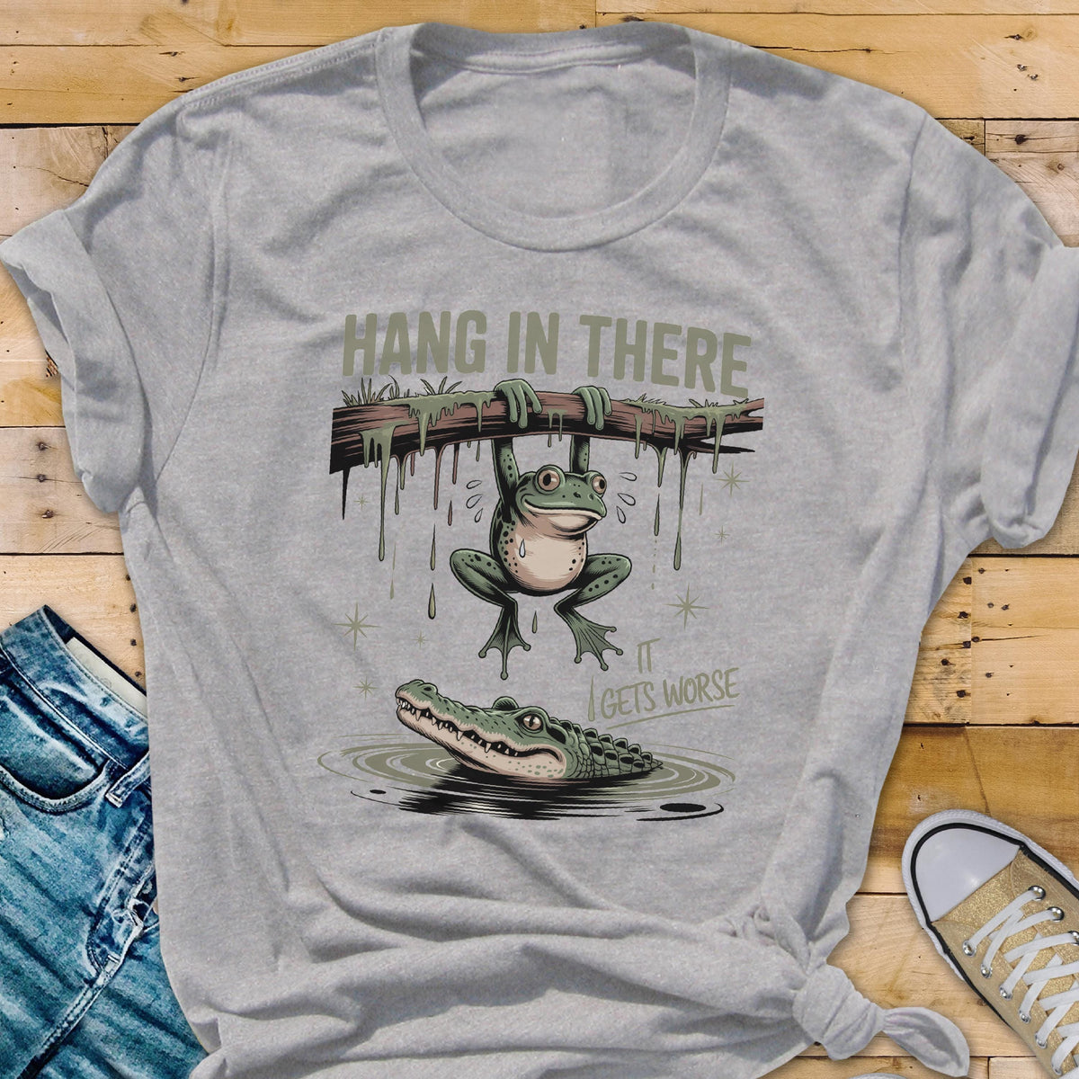 May include: A gray t-shirt with a humorous graphic. The design features a frog hanging from a branch above an alligator in water, with the text "HANG IN THERE" and "IT GETS WORSE".