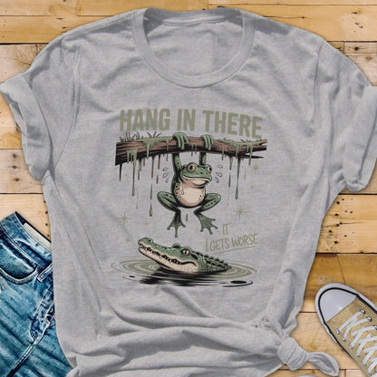 May include: A gray t-shirt with a humorous graphic. The design features a frog hanging from a branch above an alligator in water, with the text "HANG IN THERE" and "IT GETS WORSE".