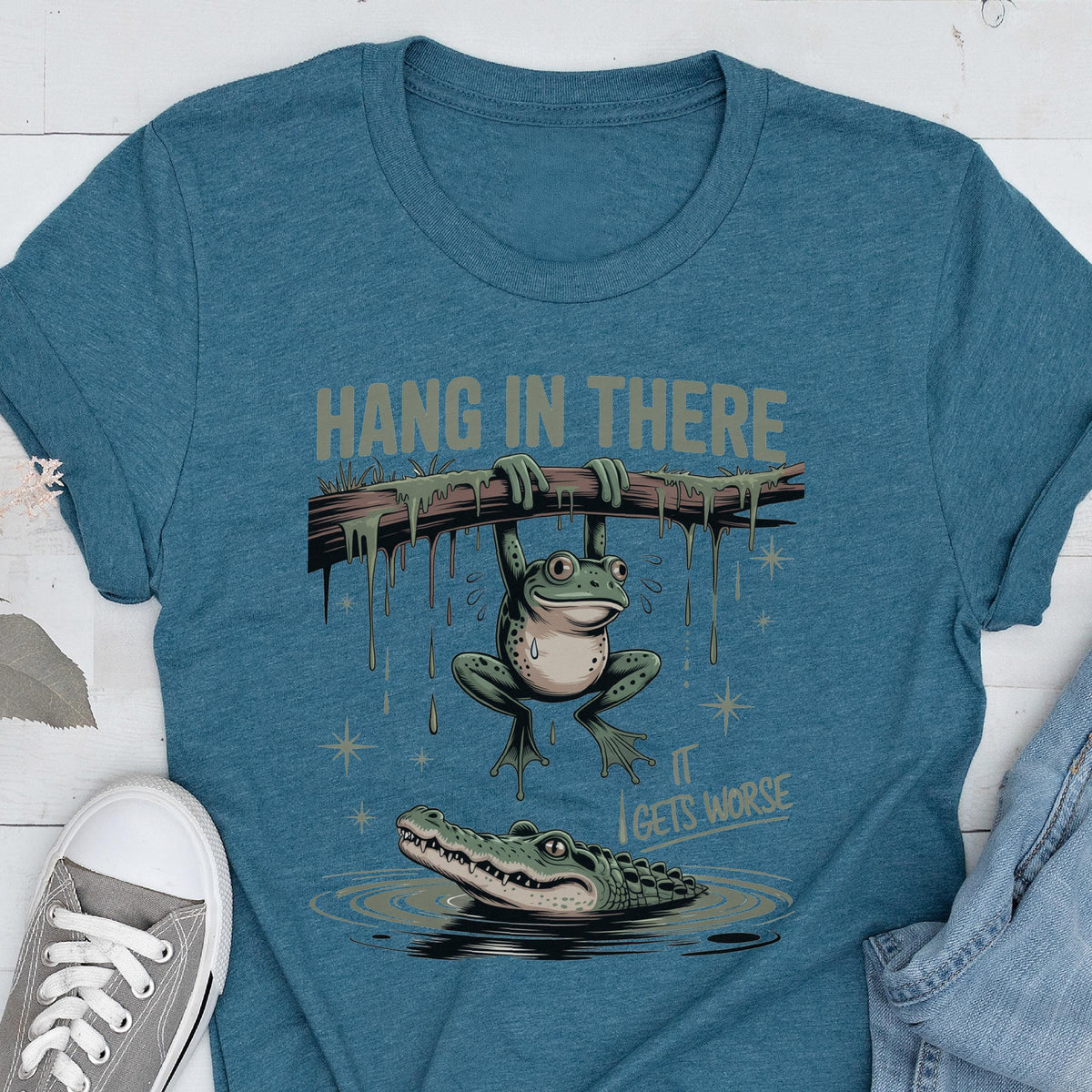 May include: Teal t-shirt with a humorous graphic of a frog hanging from a branch above an alligator in water. The text "HANG IN THERE" is above the frog, and "IT GETS WORSE" is below the alligator.