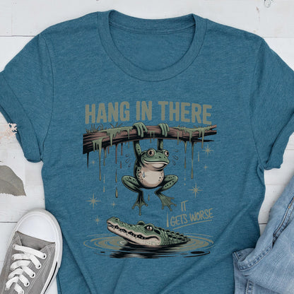 May include: Teal t-shirt with a humorous graphic of a frog hanging from a branch above an alligator in water. The text "HANG IN THERE" is above the frog, and "IT GETS WORSE" is below the alligator.