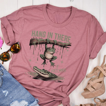 May include: Dusty rose t-shirt featuring a cartoon frog hanging from a branch with the text "HANG IN THERE." Below, an alligator swims in water, with the text "IT GETS WORSE." The design is in shades of green, brown, and white.