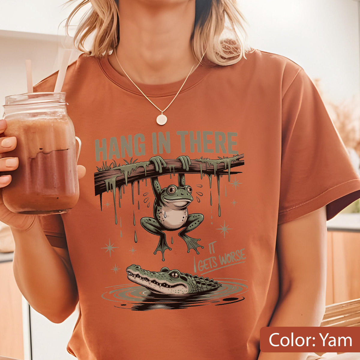 May include: Yam-colored t-shirt featuring a cartoon frog hanging from a branch above an alligator in water. The text "HANG IN THERE" is above the frog, and "IT GETS WORSE" is below. The shirt is a casual wear item.