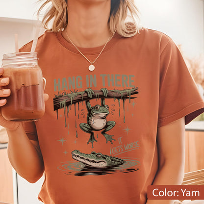 May include: Yam-colored t-shirt featuring a cartoon frog hanging from a branch above an alligator in water. The text "HANG IN THERE" is above the frog, and "IT GETS WORSE" is below. The shirt is a casual wear item.