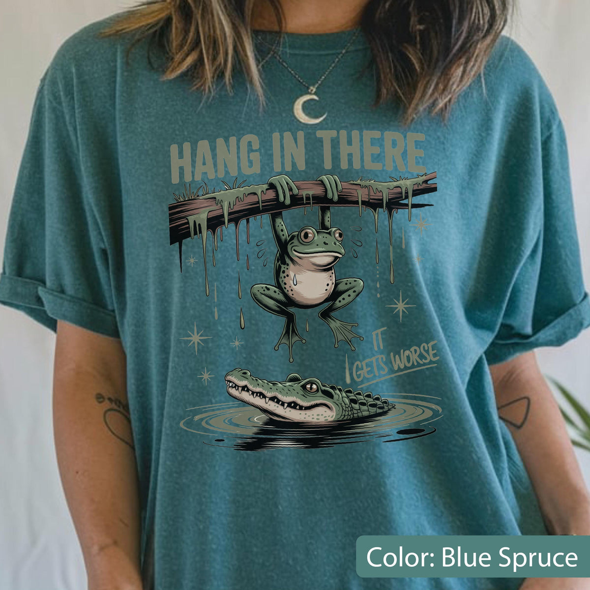May include: Blue spruce t-shirt with a humorous graphic. The design features a frog hanging from a branch above an alligator in water, with the text "Hang in There, It Gets Worse". The shirt has rolled-up sleeves.