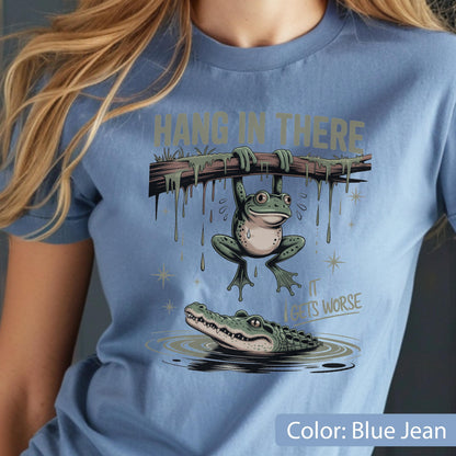 May include: Blue jean colored t-shirt with a humorous graphic. The graphic features a frog hanging from a branch above an alligator in water. The text "Hang in there" is above the frog, and "It gets worse" is below.