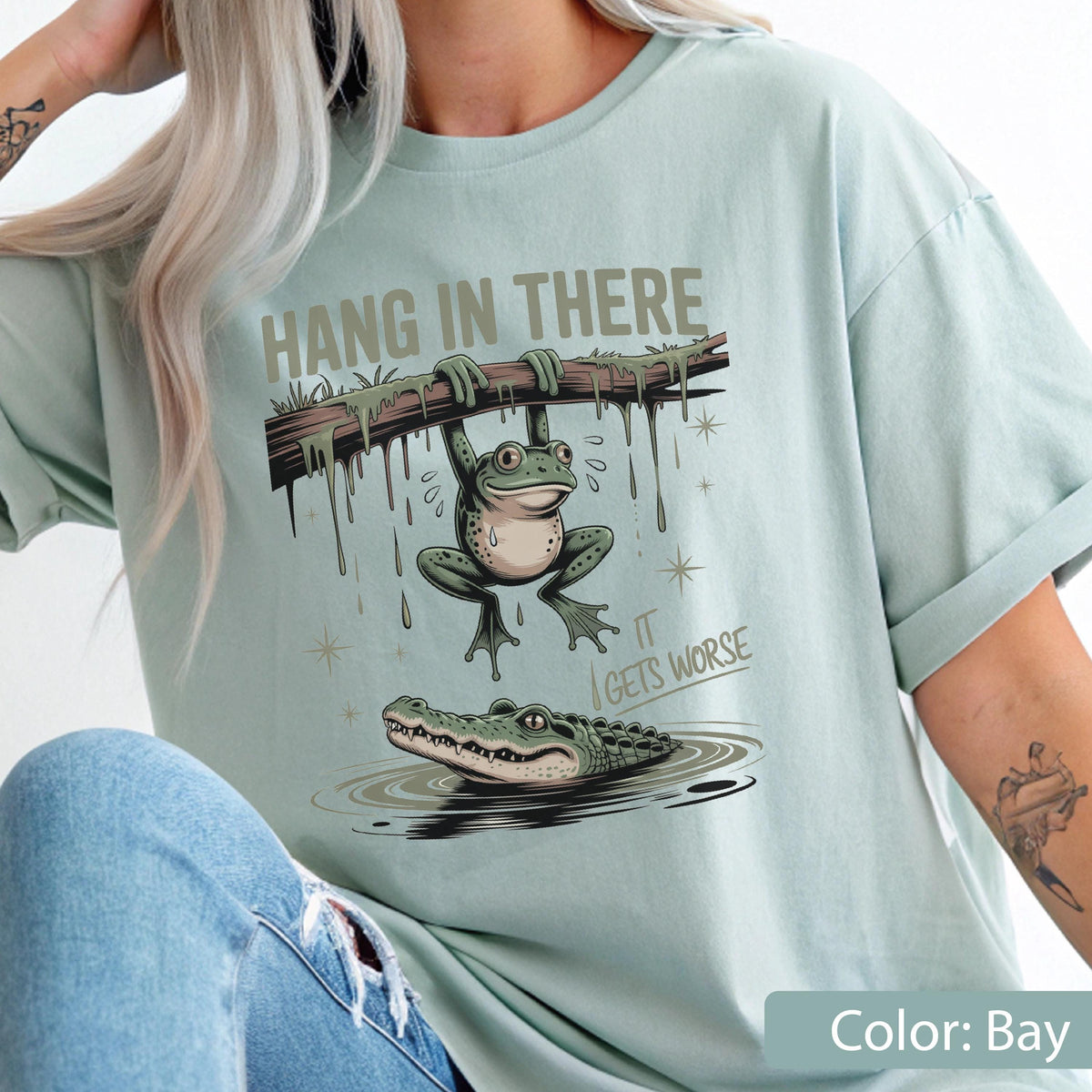 May include: Bay-colored t-shirt featuring a humorous illustration. A frog hangs from a branch above an alligator in water. The text reads "HANG IN THERE" and "IT GETS WORSE". The shirt is a relaxed fit.