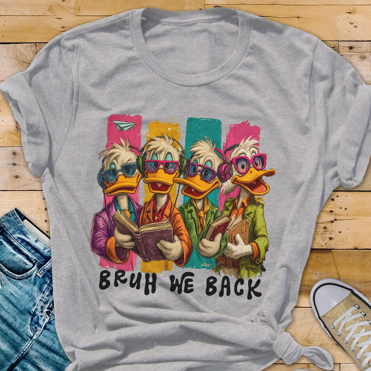May include: A light gray t-shirt featuring a cartoon image of four ducks wearing glasses and holding books. The ducks are standing in front of colorful vertical stripes. The text "BRUH WE BACK" is printed below the ducks.