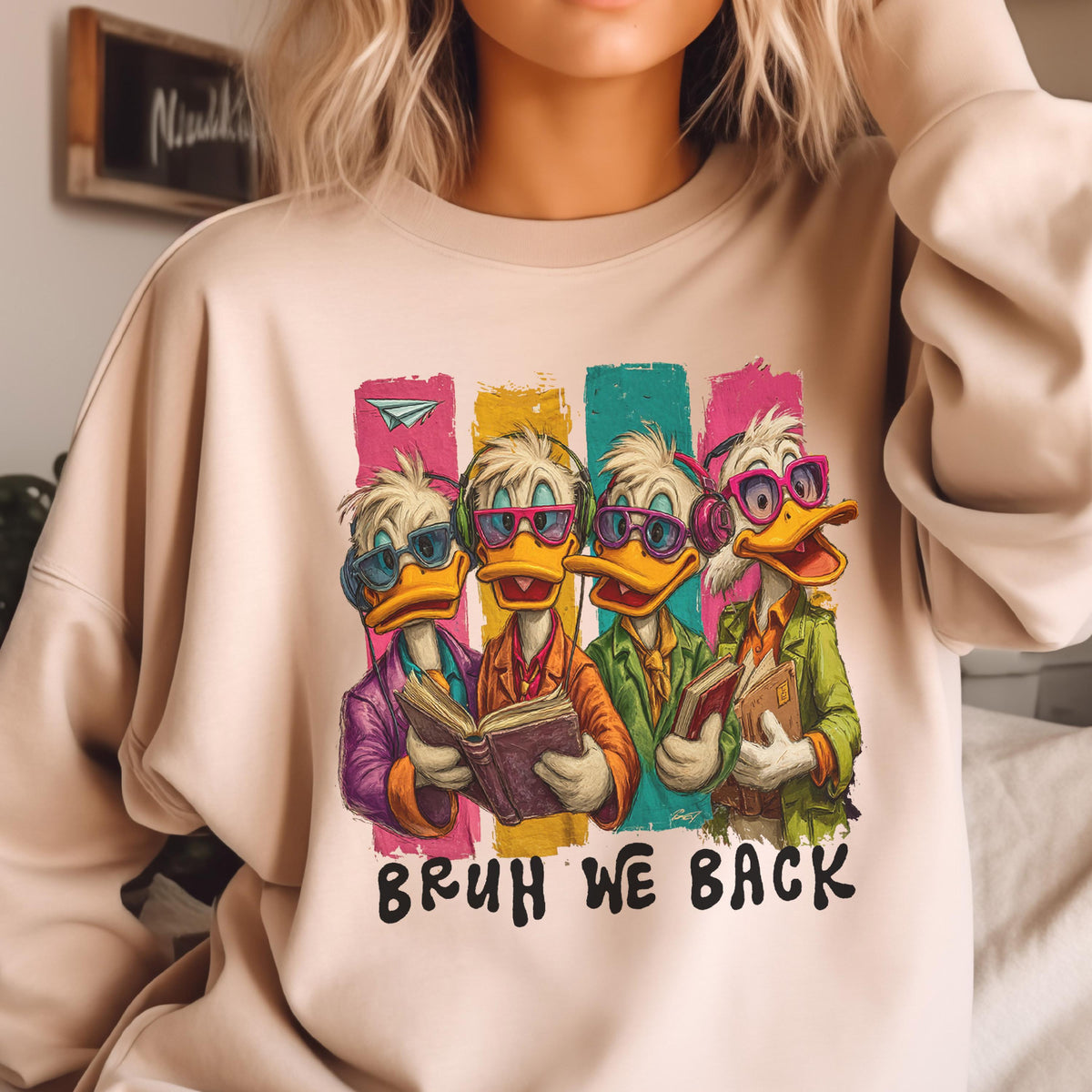 May include: Beige sweatshirt featuring a cartoon graphic of four ducks wearing sunglasses and headphones, each holding a book. The ducks are set against colorful vertical stripes, with the text "BRUH WE BACK" below.