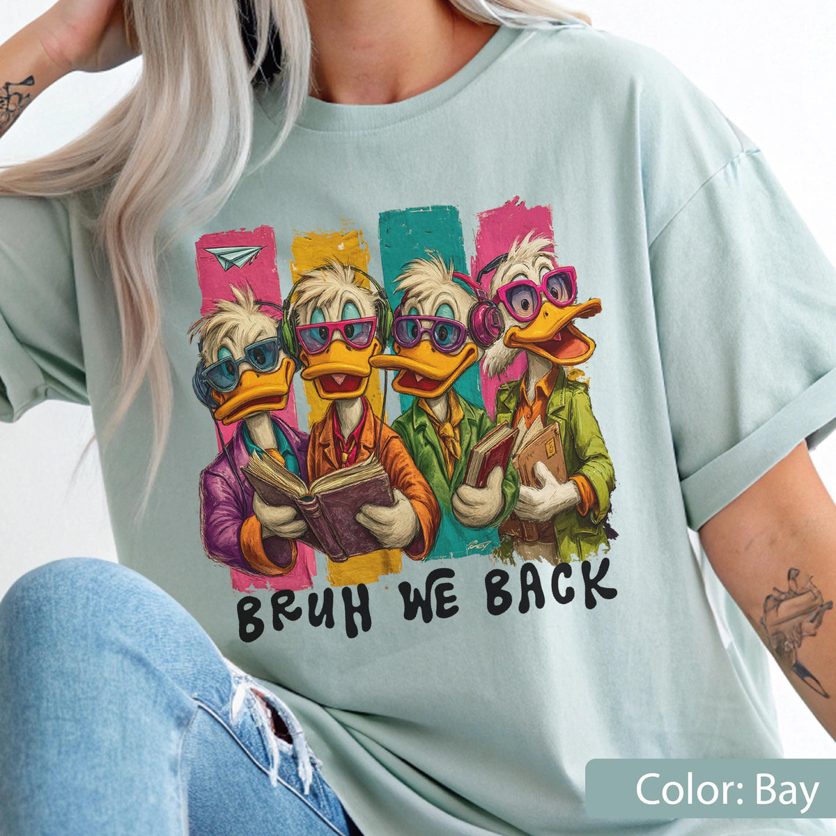 May include: A light blue t-shirt featuring a cartoon graphic of four ducks wearing glasses and holding books. The ducks are set against colorful vertical stripes, with the text "BRUH WE BACK" below. The shirt is a relaxed fit.