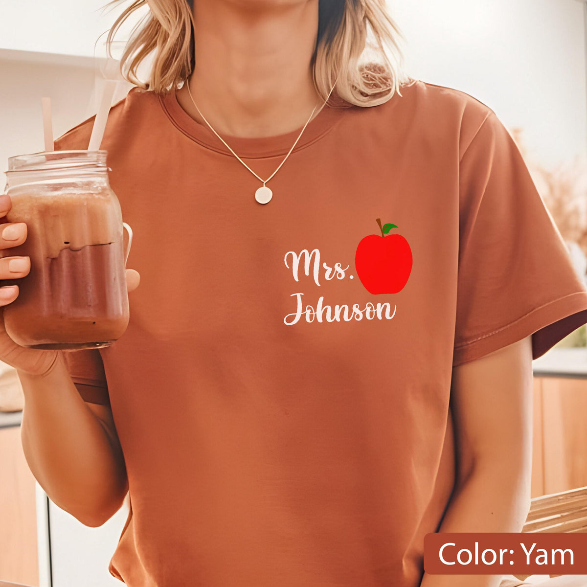 May include: A yam-colored short-sleeve t-shirt featuring a graphic on the left chest area. The graphic includes the white text "Mrs. Johnson" written in a cursive script font next to a bright red apple illustration with a small green leaf.