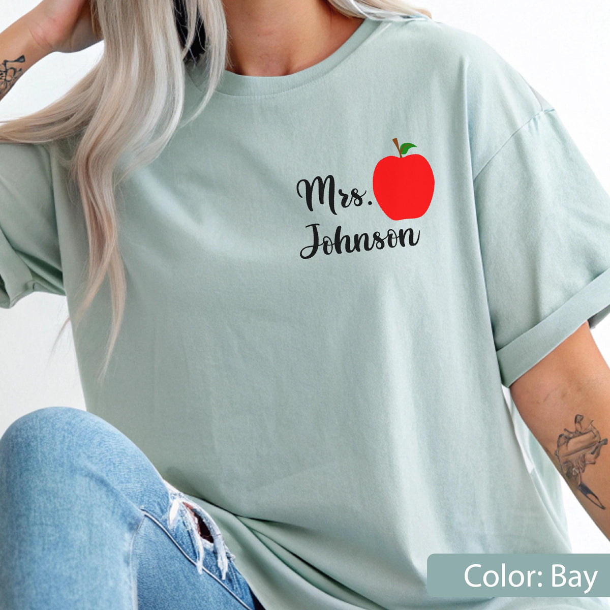 May include: A light sage green short-sleeve t-shirt featuring a graphic on the upper left chest. The graphic consists of a bright red apple with a green leaf and the black cursive text Mrs. Johnson positioned directly below the apple.