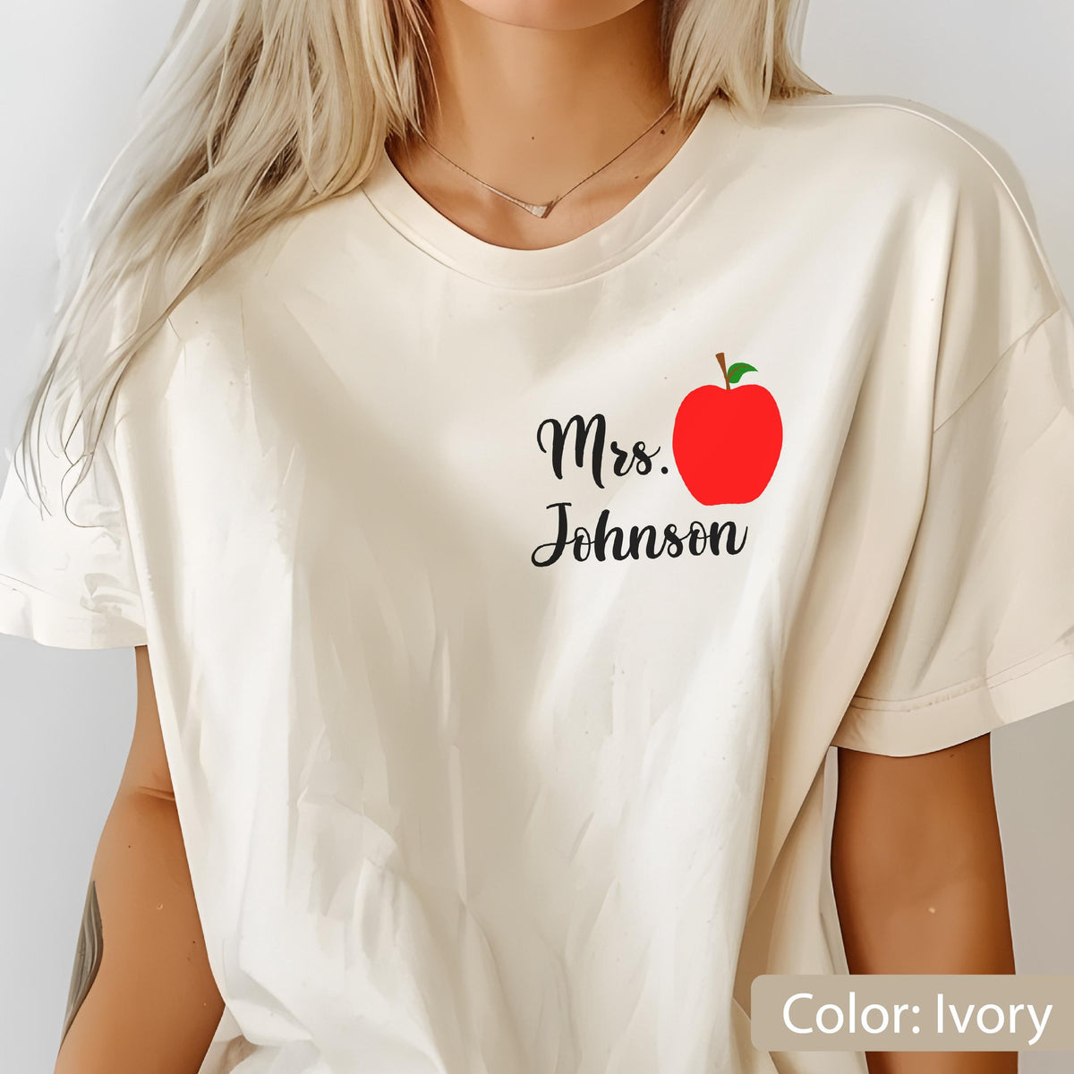May include: An ivory t-shirt featuring a graphic on the chest with the text Mrs. Johnson in black cursive script next to a bright red apple illustration with a green leaf. The shirt has a crew neckline and short sleeves in a relaxed fit.