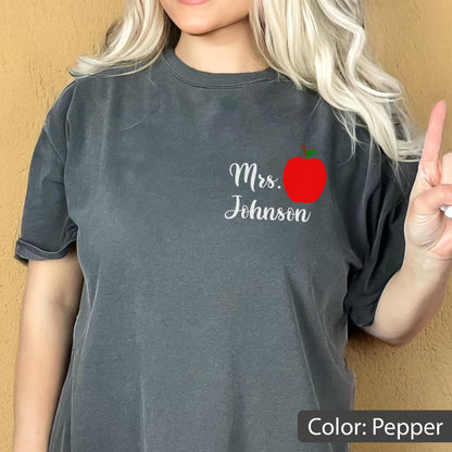 May include: A charcoal gray short-sleeve t-shirt featuring a graphic on the left chest. The graphic includes a bright red apple illustration positioned above the white cursive text that reads Mrs. Johnson. The shirt has a crew neckline.