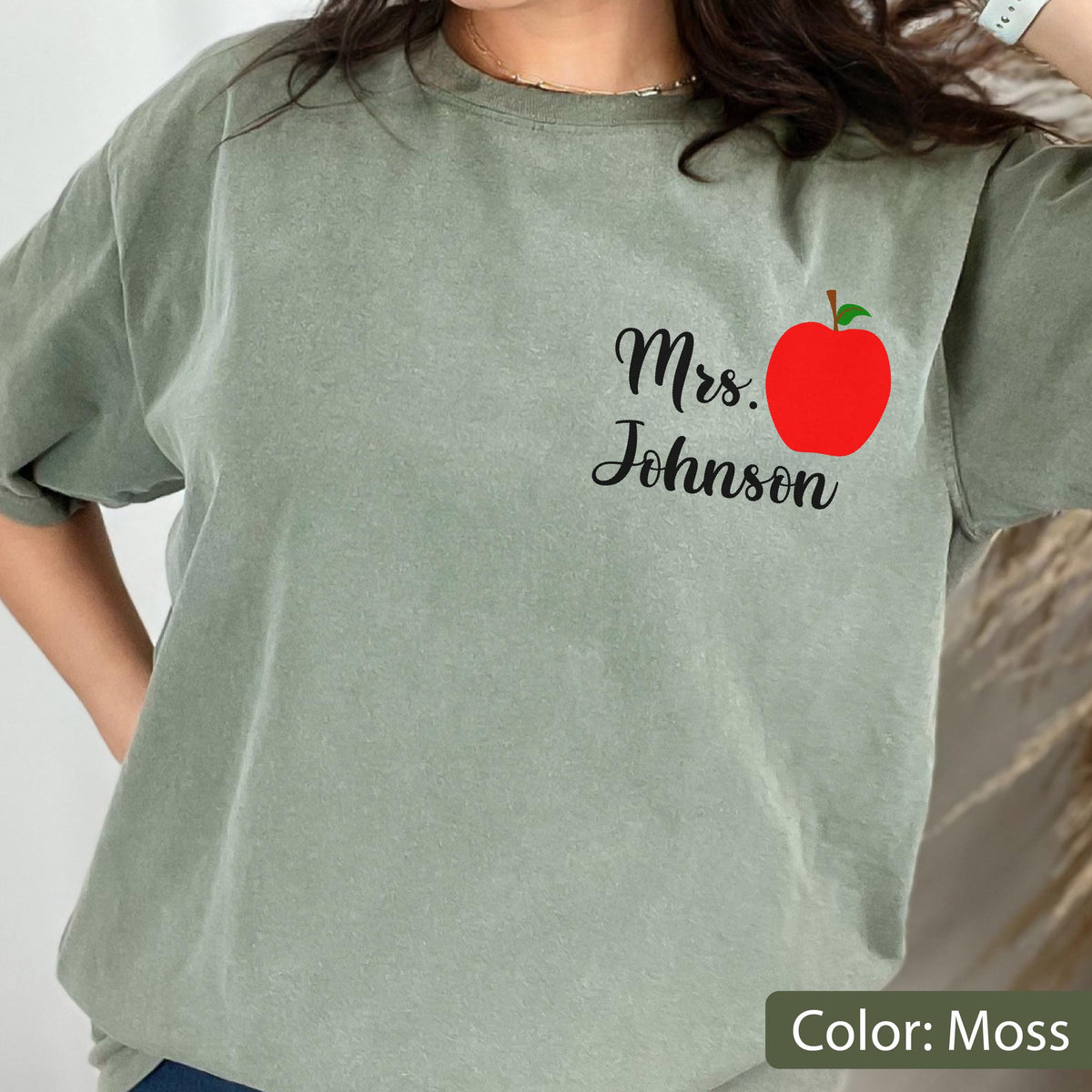 May include: A moss green short-sleeve t-shirt featuring a graphic on the upper left chest. The graphic consists of a bright red apple illustration positioned next to the black cursive text that reads Mrs. Johnson in a clean, legible font style.